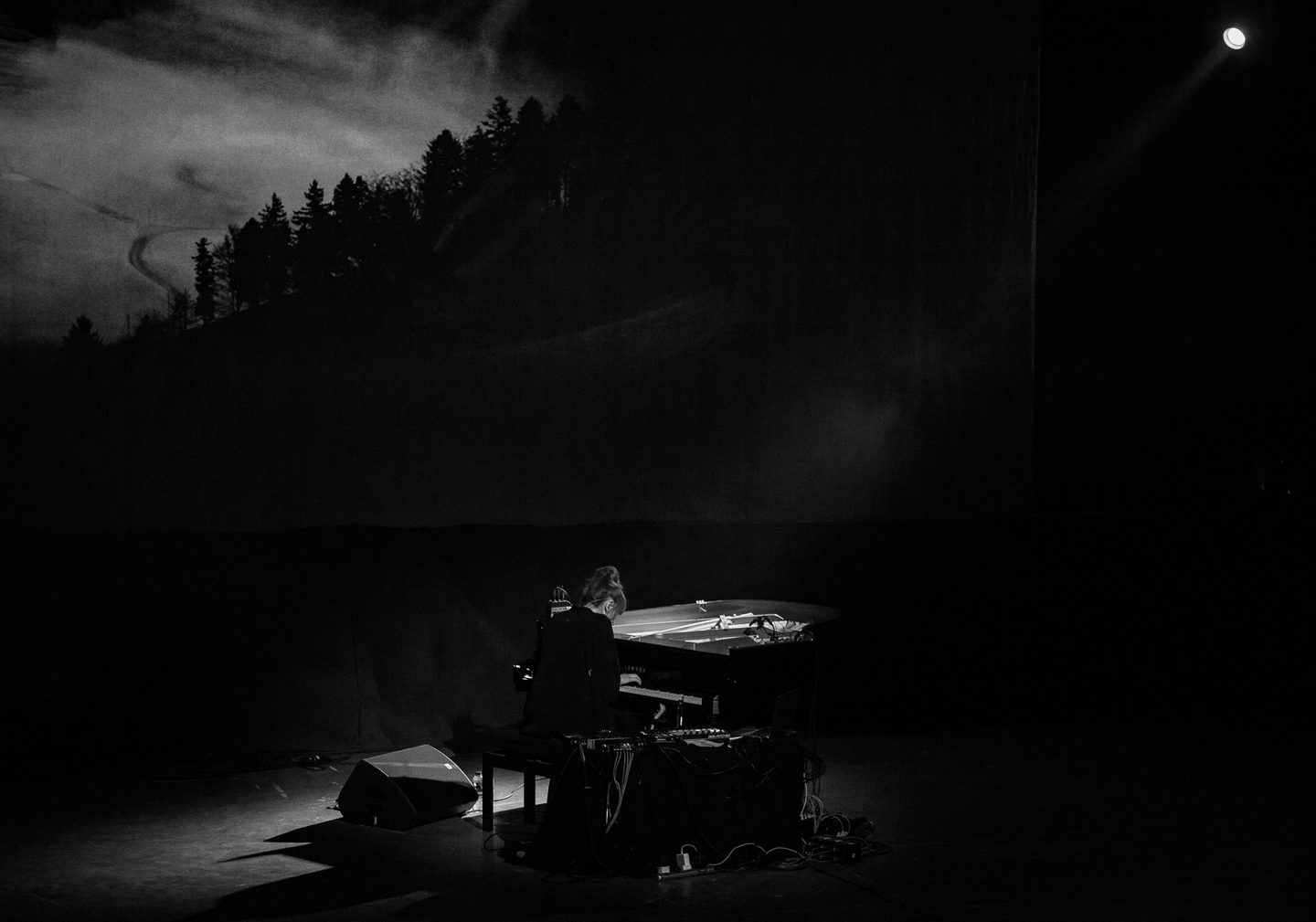 A black‑and‑white photograph of a musician seated at a piano on a dimly lit stage. The performer is shown from behind, illuminated by a soft overhead light. Cables and equipment surround the piano. In the background, a large screen displays a dark, atmospheric image of a forested hillside under cloudy skies, creating a moody and cinematic setting.