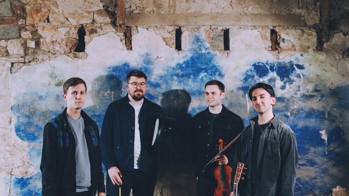 Four men stand against a weathered wall, holding musical instruments.