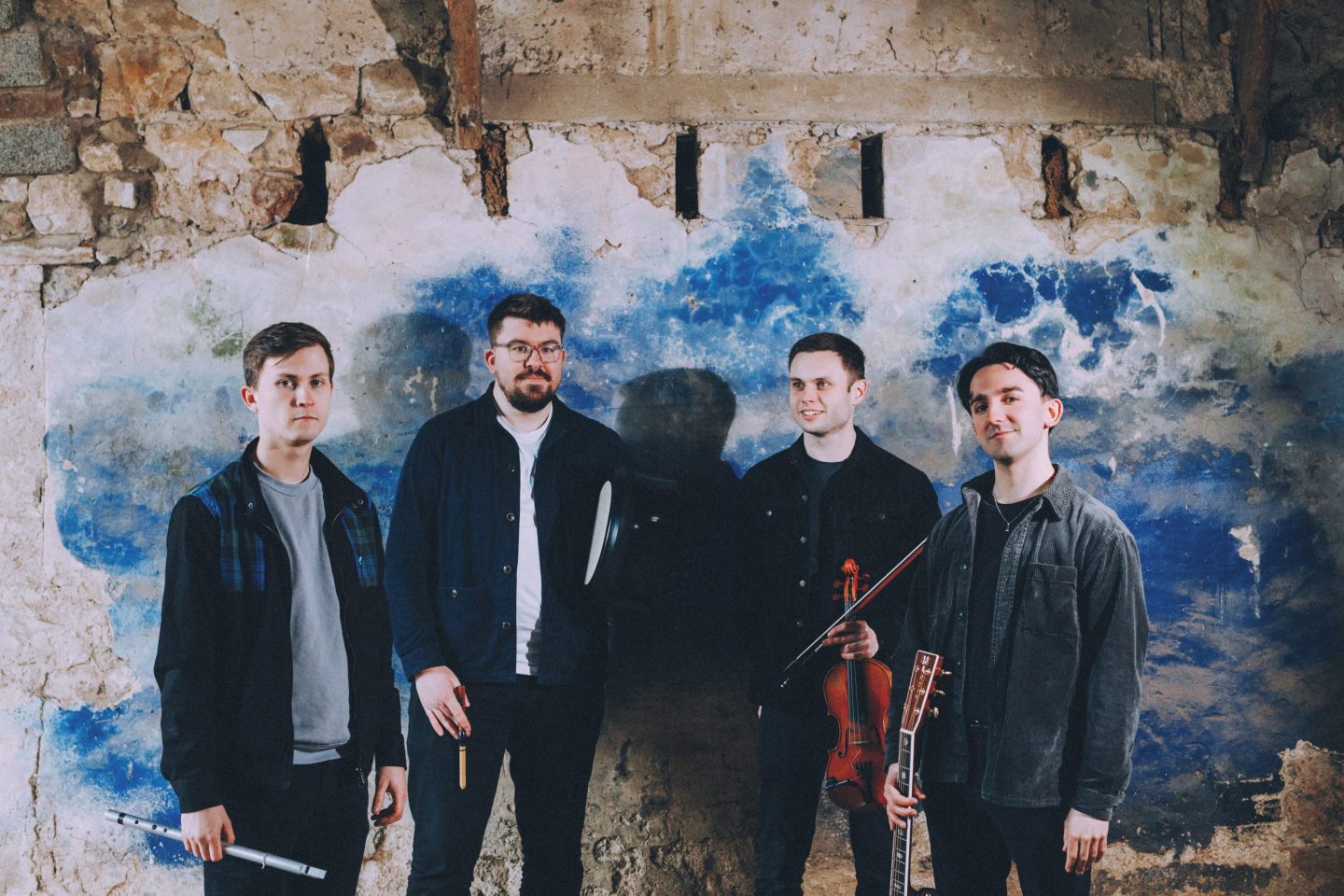 Four men stand against a weathered wall, holding musical instruments.