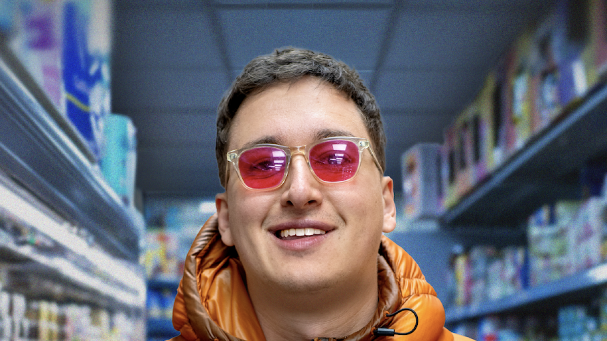 Smiling person wearing pink sunglasses and an orange puffer jacket, standing in a brightly lit supermarket aisle.