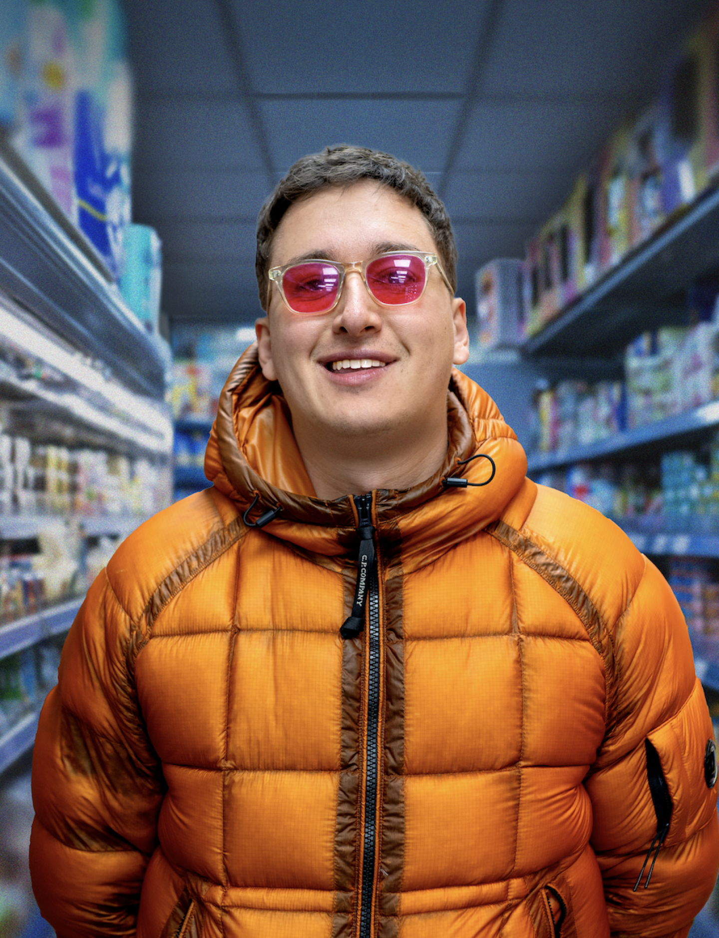 Smiling person wearing pink sunglasses and an orange puffer jacket, standing in a brightly lit supermarket aisle.