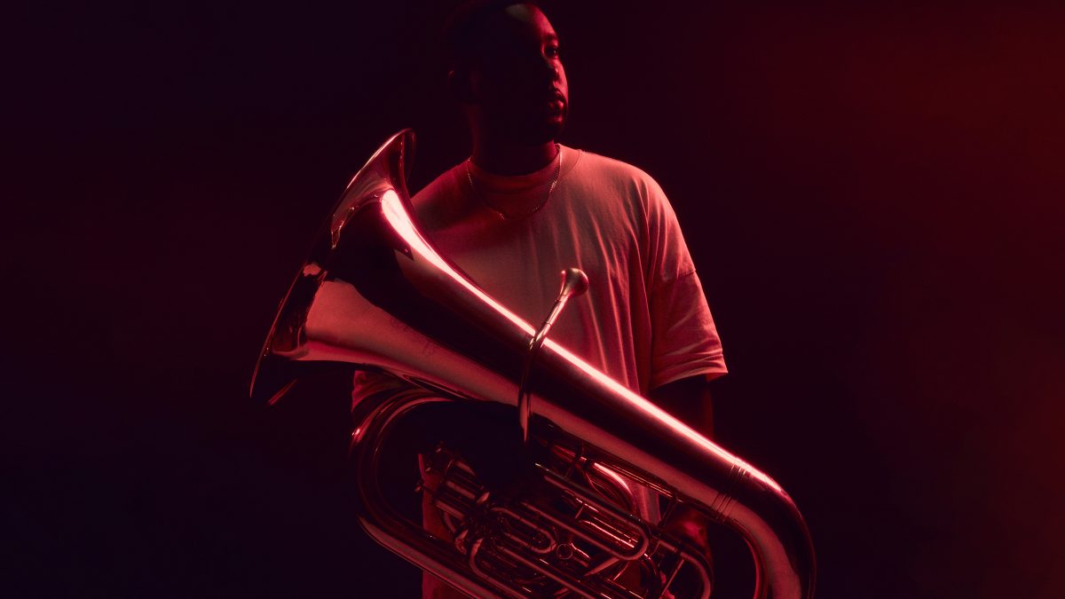 A person standing in deep red lighting while holding a large brass tuba. The reflective surface of the instrument catches the red light, creating dramatic highlights. The individual wears a light-colored shirt, and the background fades into dark shadows.