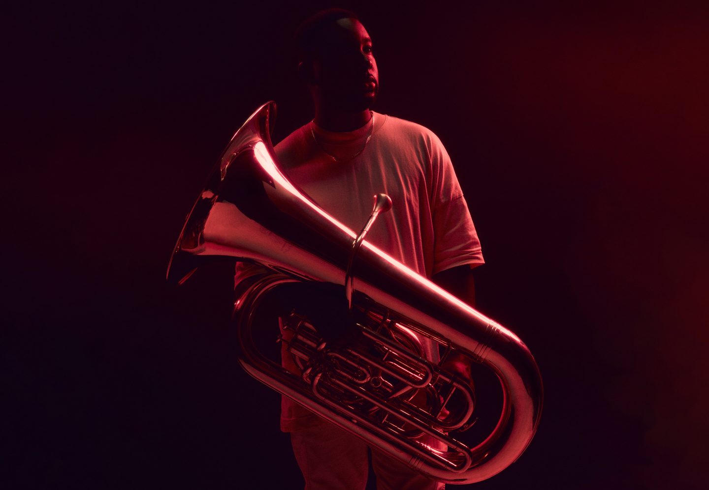 A person standing in deep red lighting while holding a large brass tuba. The reflective surface of the instrument catches the red light, creating dramatic highlights. The individual wears a light-colored shirt, and the background fades into dark shadows.