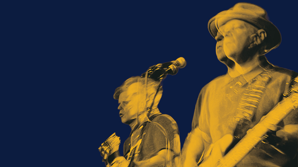 Two musicians playing guitars on stage, shown in yellow tones against a dark blue background with motion blur.