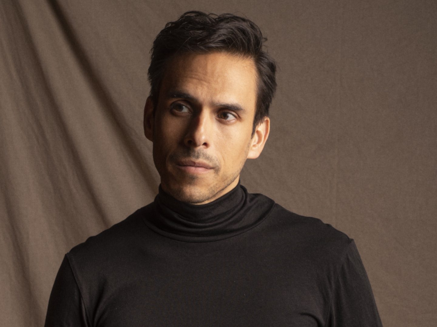 A man wearing a black polo neck stands against a brown background
