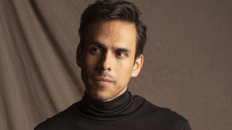 A man wearing a black polo neck stands against a brown background