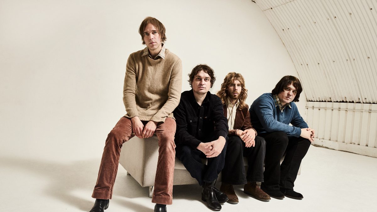 Four individuals sit in a row on a low platform in a bright, white studio space with a curved wall behind them. All are seated facing forward, dressed in casual clothing.