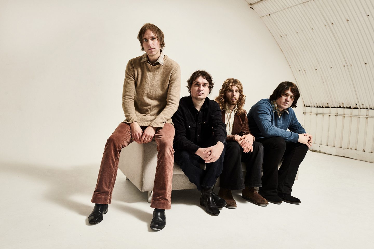 Four individuals sit in a row on a low platform in a bright, white studio space with a curved wall behind them. All are seated facing forward, dressed in casual clothing.