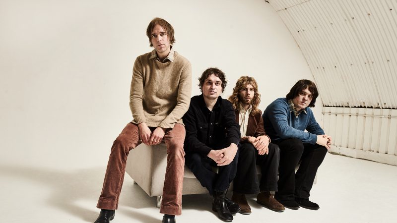 Four individuals sit in a row on a low platform in a bright, white studio space with a curved wall behind them. All are seated facing forward, dressed in casual clothing.