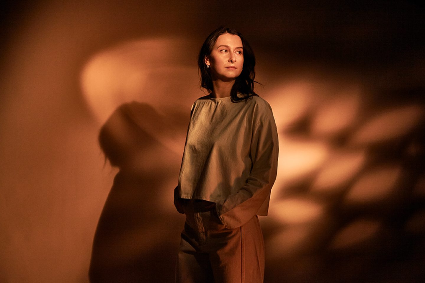 A figure with long brown hair looking over one should, stood against a terracotta background of dappled sunlight.