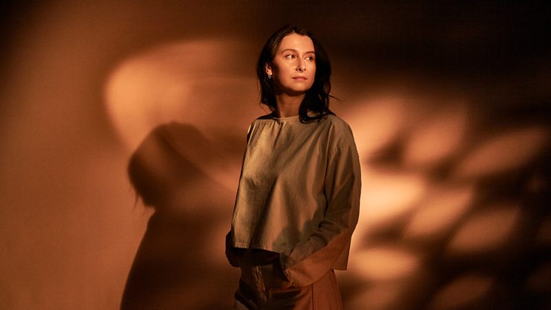 A figure with long brown hair looking over one should, stood against a terracotta background of dappled sunlight.