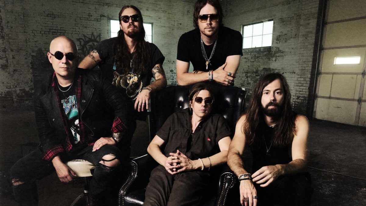 Five individuals pose together in a dim, industrial-style room, with one seated in a large chair and the others standing or sitting around it.