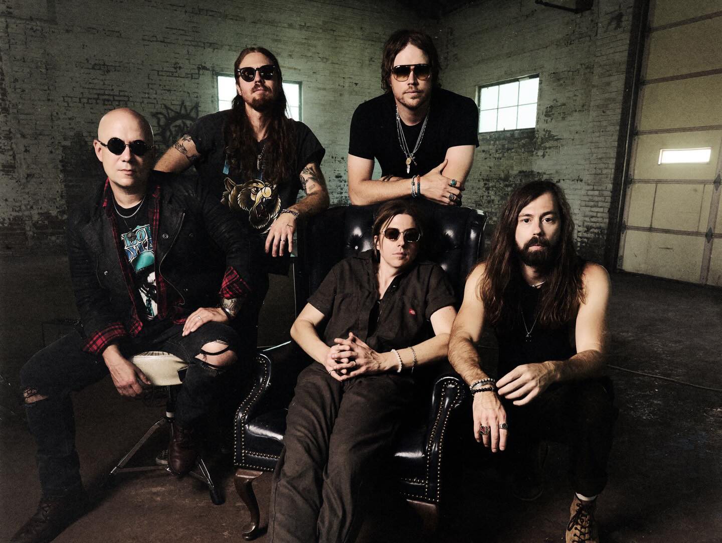 Five individuals pose together in a dim, industrial-style room, with one seated in a large chair and the others standing or sitting around it.