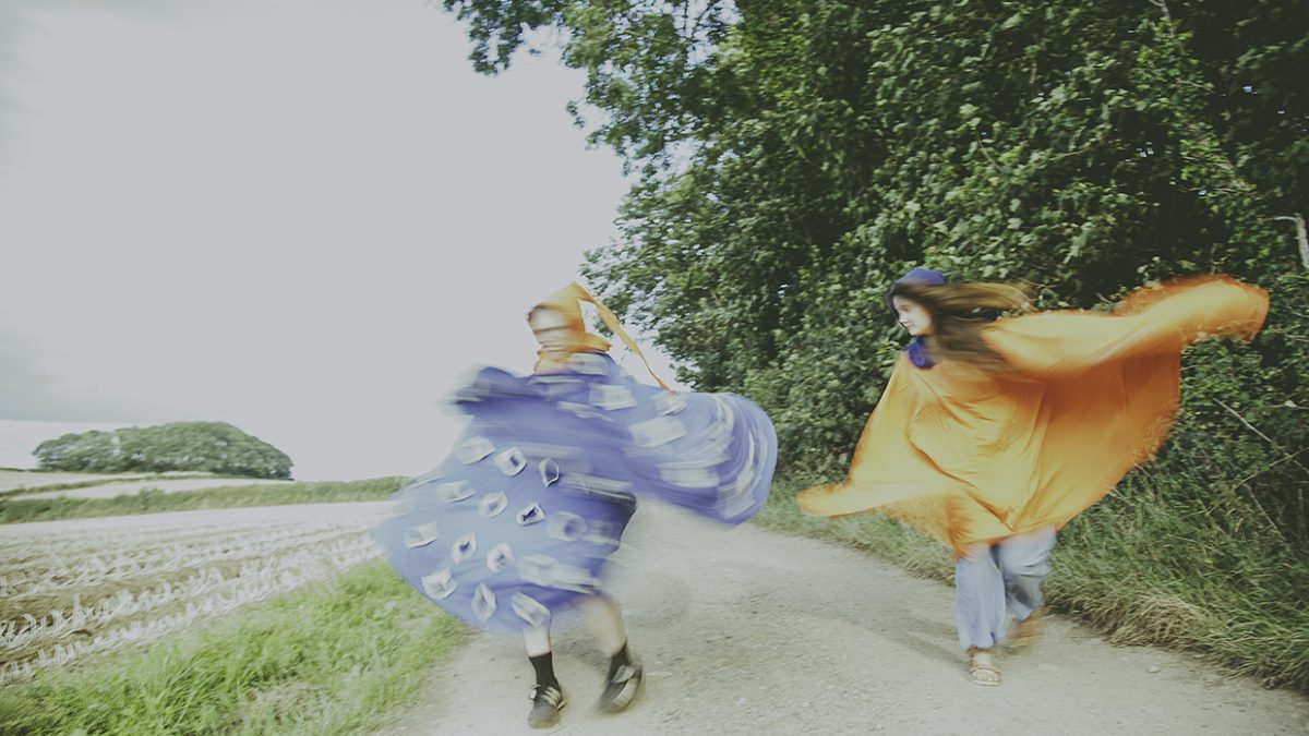 Two people are moving energetically along a rural dirt path bordered by tall green trees on one side and an open field on the other. Their motion creates a blurred effect. One person is wrapped in or wearing a billowing bright blue fabric decorated with circular patterns, while the other is wrapped in bright orange fabric that flows outward as they move. The scene has a soft, slightly overexposed look, and the path curves gently through the landscape under a pale sky.