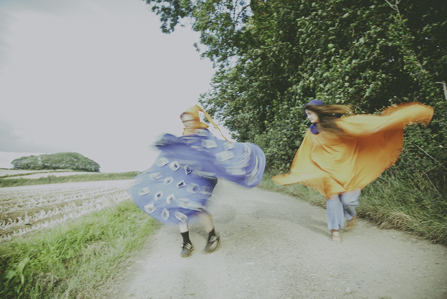 Two people are moving energetically along a rural dirt path bordered by tall green trees on one side and an open field on the other. Their motion creates a blurred effect. One person is wrapped in or wearing a billowing bright blue fabric decorated with circular patterns, while the other is wrapped in bright orange fabric that flows outward as they move. The scene has a soft, slightly overexposed look, and the path curves gently through the landscape under a pale sky.