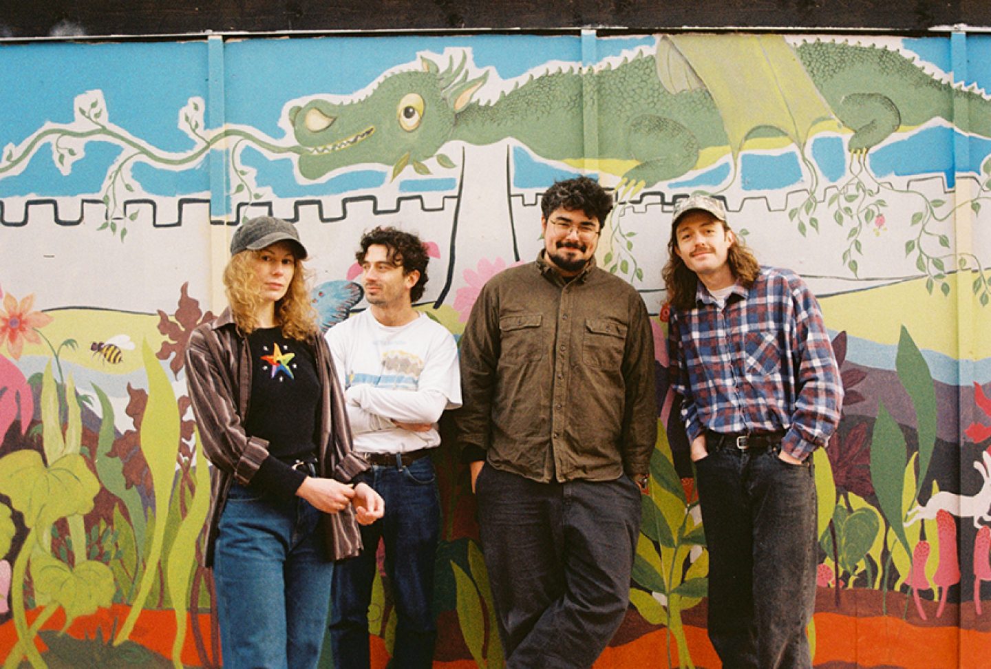 Four individuals stand in front of a colorful mural featuring a green dragon above a castle wall and plants below.