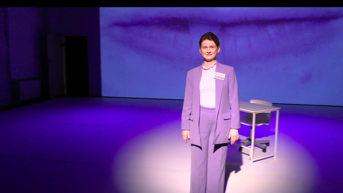A person stands centered on a dimly lit stage, illuminated by a circular spotlight. They are wearing a light purple suit with a name tag on the chest and are positioned in front of a plain desk and chair. The background features a large projected close-up image of a person's lips in soft blue tones, filling the wall behind them. The overall lighting is cool and theatrical, creating a minimalist performance setting.