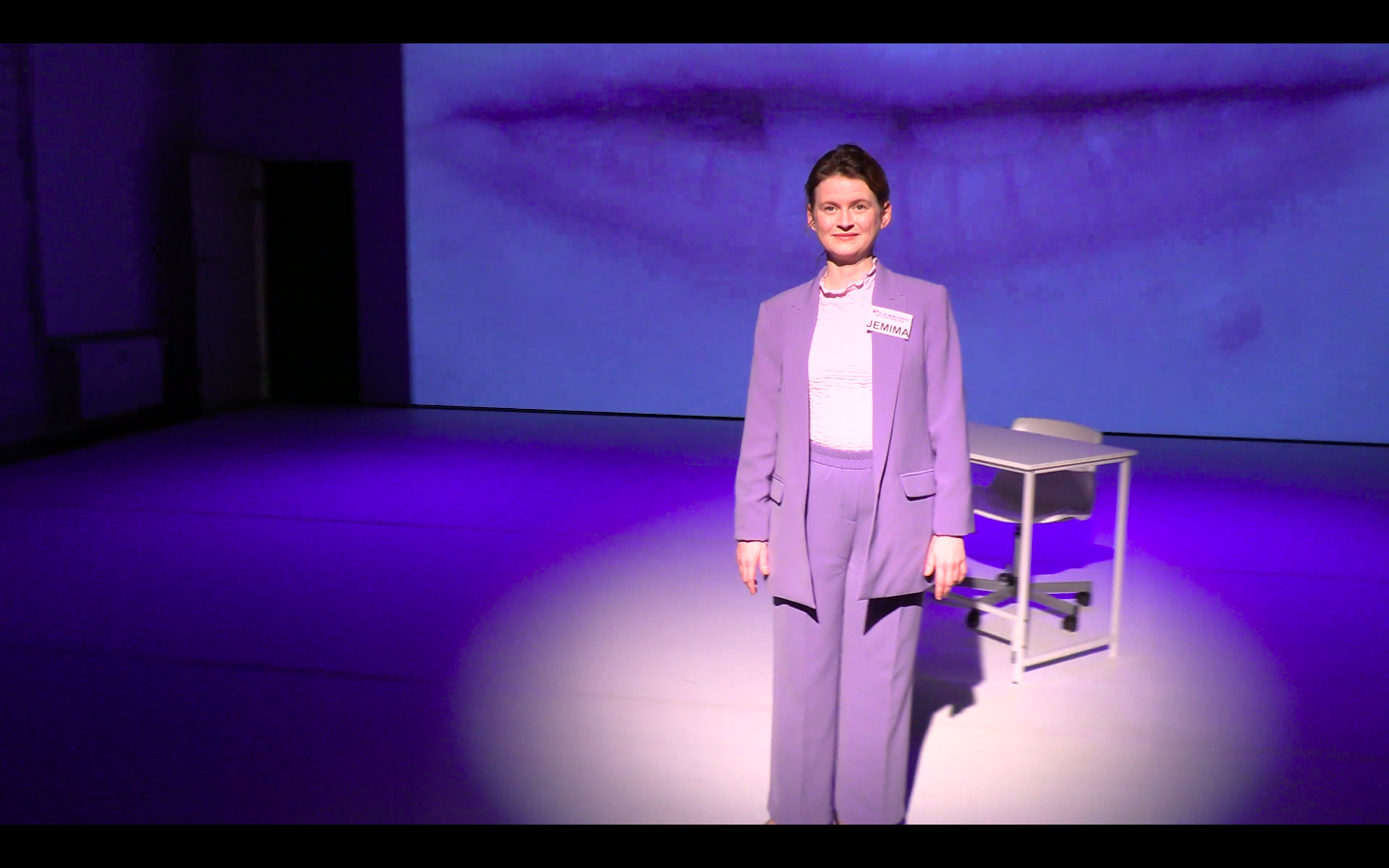 A person stands centered on a dimly lit stage, illuminated by a circular spotlight. They are wearing a light purple suit with a name tag on the chest and are positioned in front of a plain desk and chair. The background features a large projected close-up image of a person's lips in soft blue tones, filling the wall behind them. The overall lighting is cool and theatrical, creating a minimalist performance setting.
