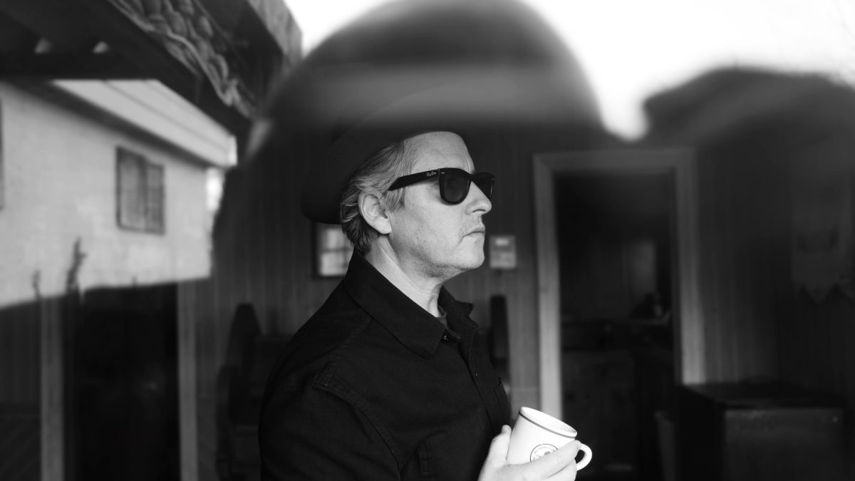 A black-and-white photo of a man in sunglasses holding a cup, standing in profile on a patio.