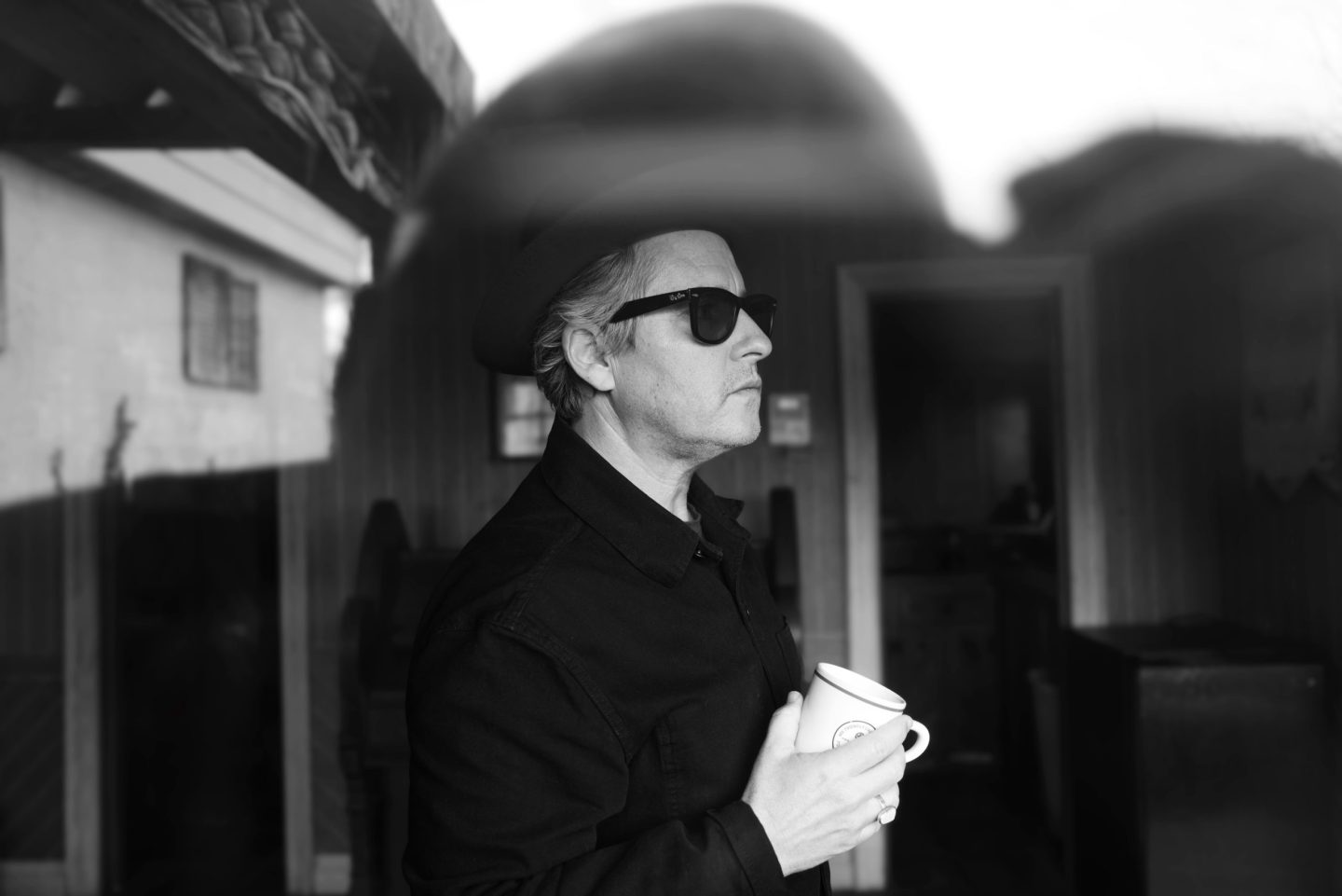 A black-and-white photo of a man in sunglasses holding a cup, standing in profile on a patio.