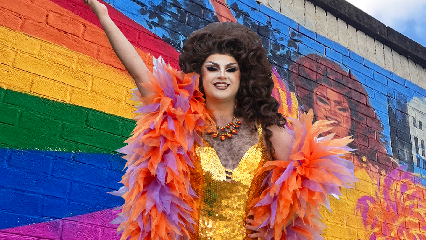 Drag performer in a gold outfit and feathered boa posing in front of a rainbow mural.