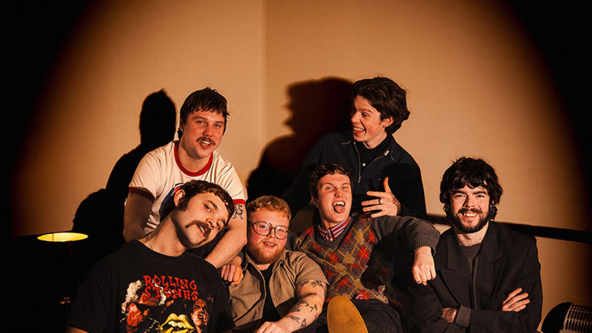 A group of six people sit close together on a couch under warm spotlight-style lighting, posing casually with relaxed, playful expressions and varied casual clothing.