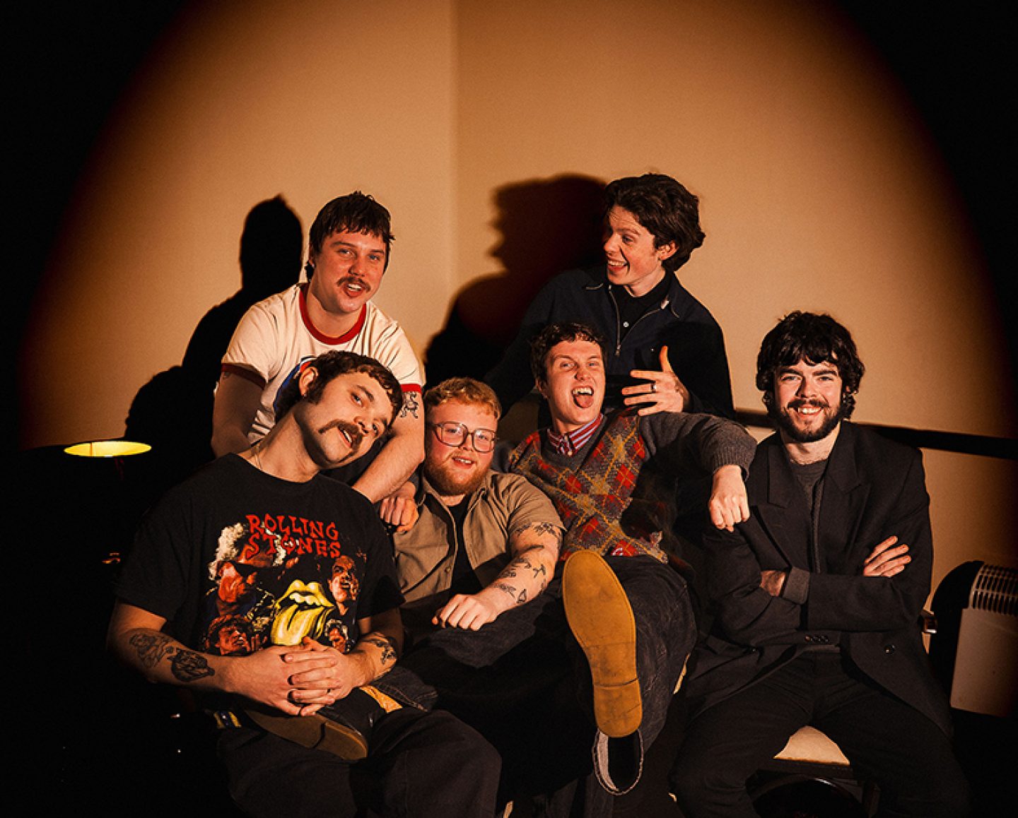 A group of six people sit close together on a couch under warm spotlight-style lighting, posing casually with relaxed, playful expressions and varied casual clothing.