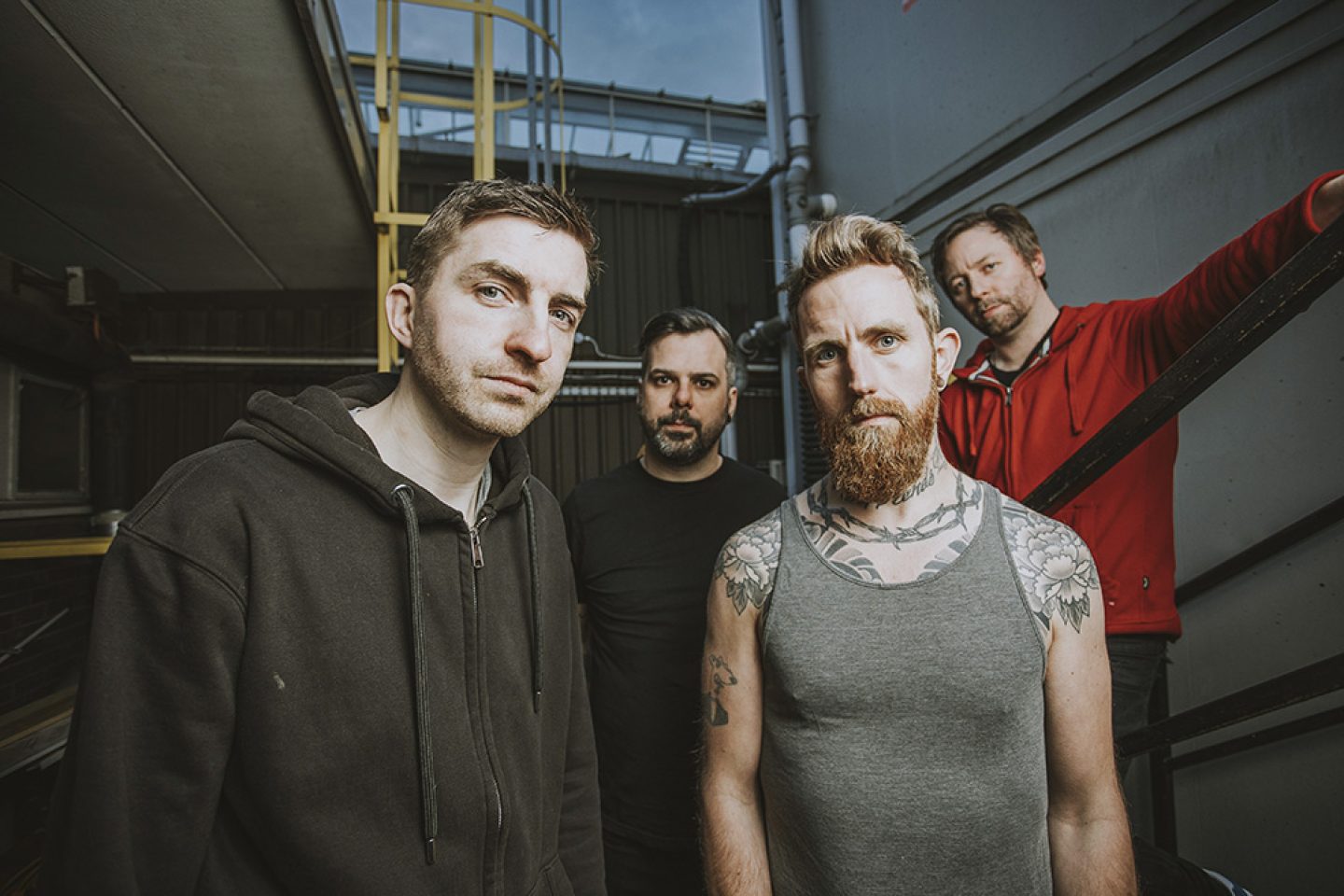 Four people stand closely together in a narrow, industrial outdoor corridor with metal railings and a yellow ladder behind them. Three face the camera in the foreground, while one leans on a railing in the background wearing a red hoodie.
