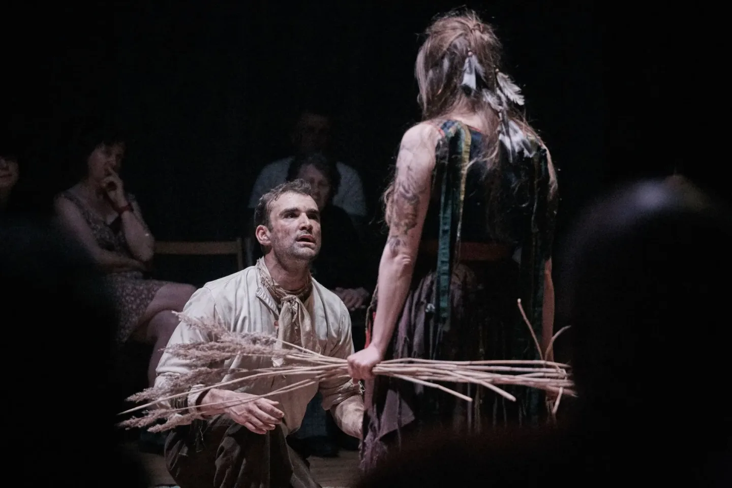 Man kneeling on a stage looks up at a woman holding a bundle of dried plants while audience members sit in the dim background.