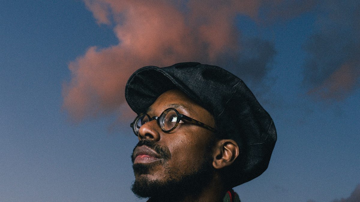 A person is standing outdoors during dusk, with a dramatic sky of deep blue tones and softly illuminated pink clouds behind them. The person is wearing a dark flat cap and a long, knitted scarf featuring red and green stripes. They are dressed in a dark coat or jacket, and the lighting suggests the sun has recently set, casting subtle shadows and highlighting the silhouette against the colorful sky. The background shows low, dark clouds along the horizon, adding to the atmospheric, moody setting.