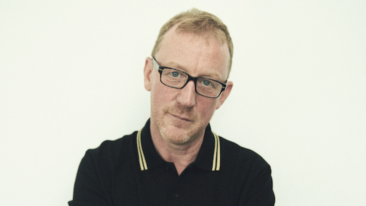 Close-up of a man with glasses wearing a black polo shirt, looking down thoughtfully against a plain light background.
