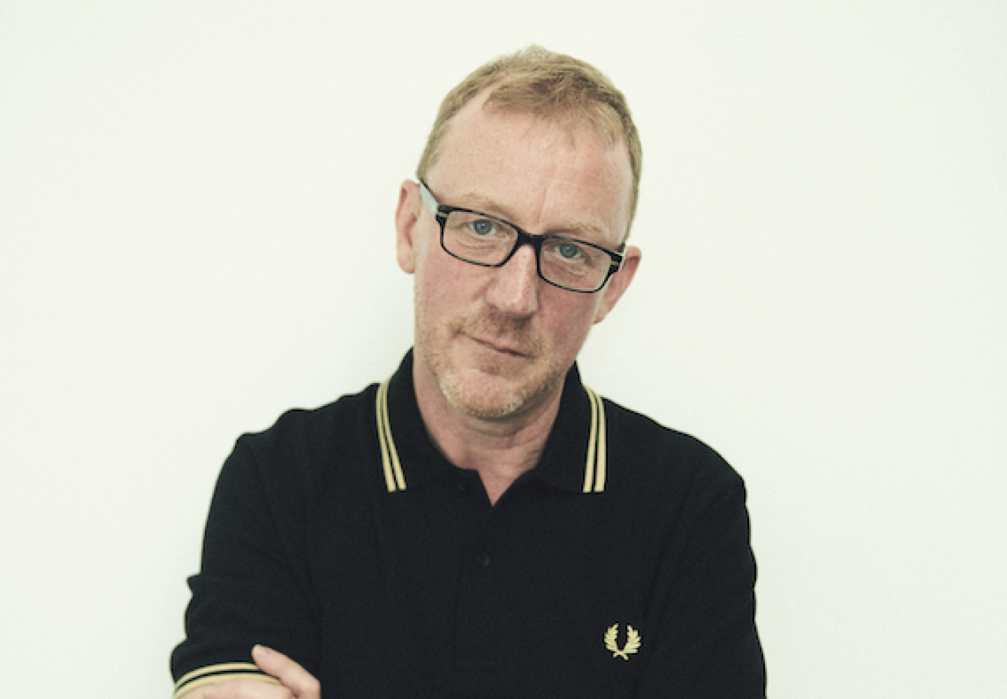 Close-up of a man with glasses wearing a black polo shirt, looking down thoughtfully against a plain light background.