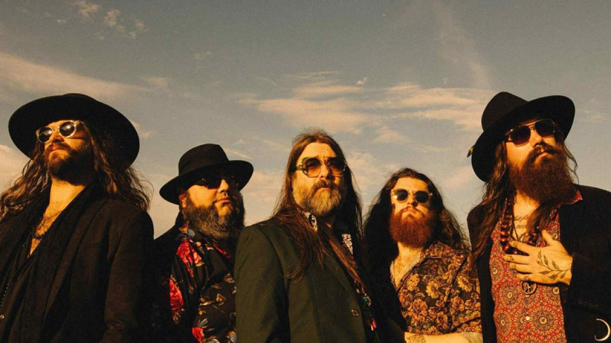 A group of five individuals standing outdoors under a partly cloudy sky, all wearing sunglasses and patterned or dark clothing, some with hats and long hair and beards, posed in a line facing slightly upward toward the light.