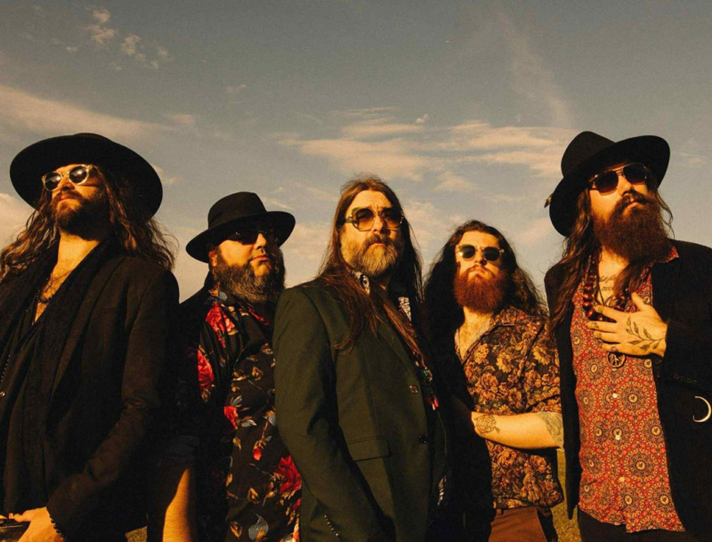 A group of five individuals standing outdoors under a partly cloudy sky, all wearing sunglasses and patterned or dark clothing, some with hats and long hair and beards, posed in a line facing slightly upward toward the light.