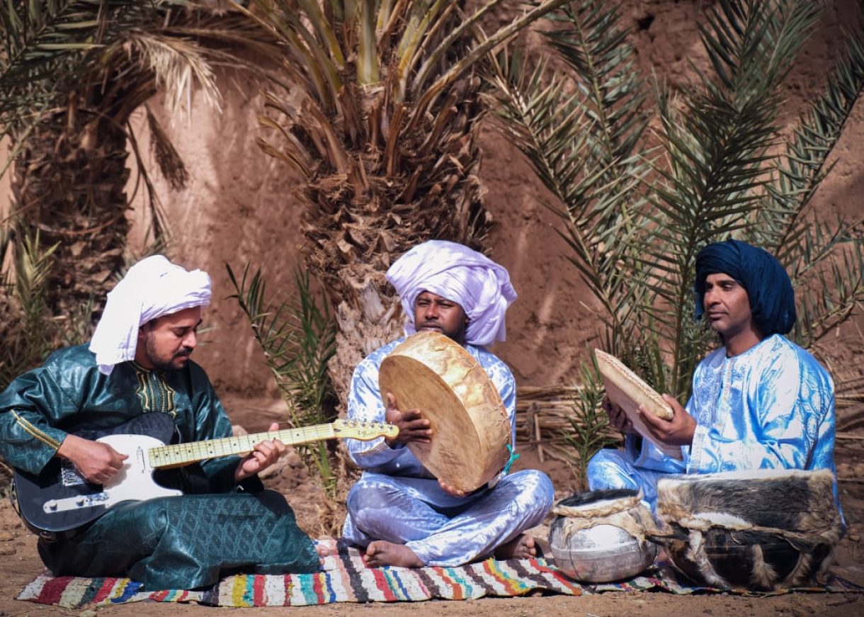 Three men in traditional clothing playing music with a guitar and drums under palm trees.