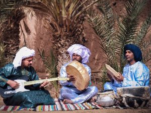 Three men in traditional clothing playing music with a guitar and drums under palm trees.