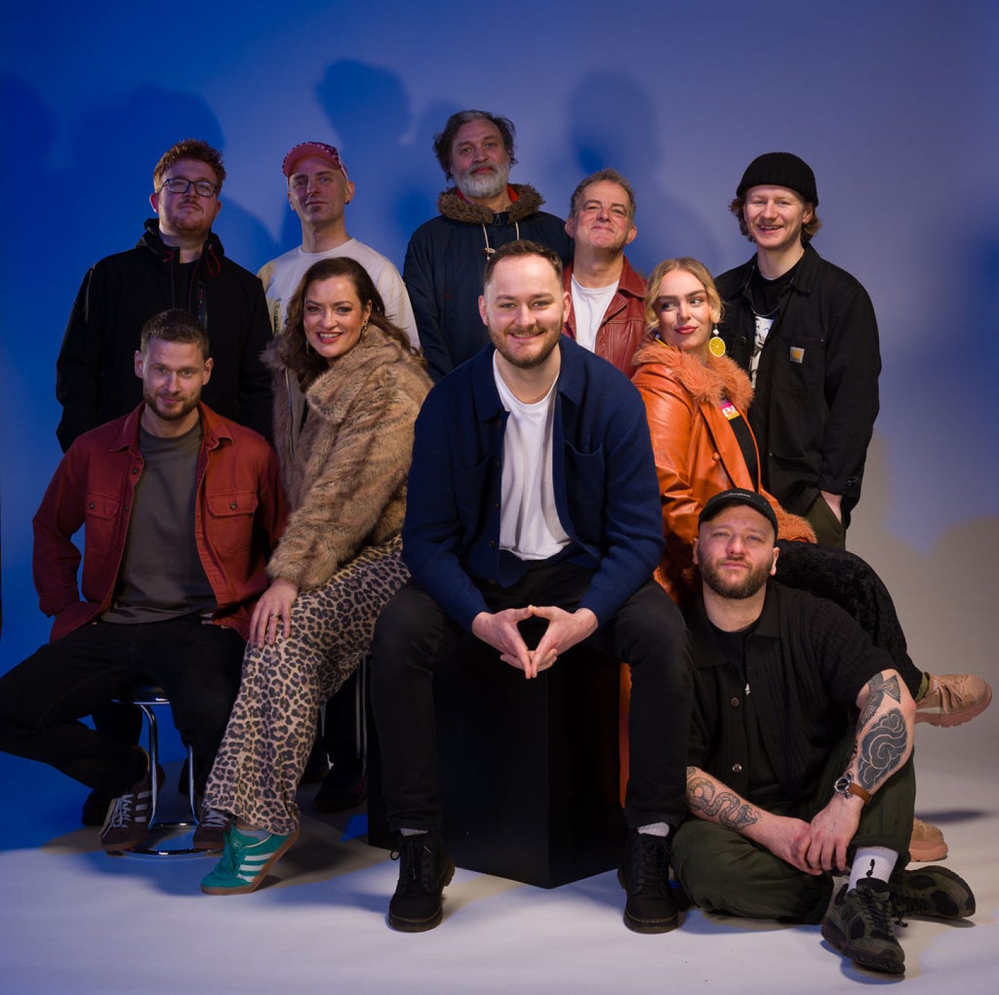 A group of ten adults pose together in a studio with a plain blue-lit background. They are arranged in two rows—some seated in front and others standing behind. The group is casually dressed in a mix of jackets, sweaters, and accessories like hats and glasses. One person in front sits centered on a cube, smiling with hands clasped, while others sit on stools or the floor. The lighting creates soft shadows behind them.