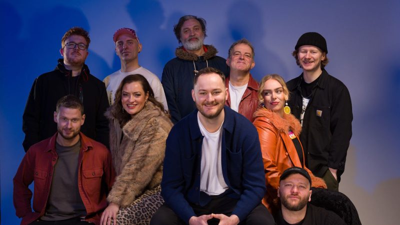 A group of ten adults pose together in a studio with a plain blue-lit background. They are arranged in two rows—some seated in front and others standing behind. The group is casually dressed in a mix of jackets, sweaters, and accessories like hats and glasses. One person in front sits centered on a cube, smiling with hands clasped, while others sit on stools or the floor. The lighting creates soft shadows behind them.