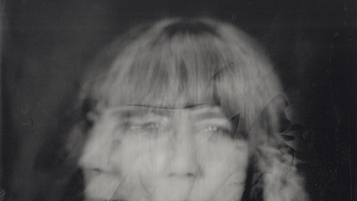 A black-and-white, heavily blurred portrait of a person with braided hair and bangs. The image appears to have multiple overlapping exposures, creating a ghostly, distorted effect where facial features are doubled and smeared. The person seems to be wearing a light-colored shirt with a collar, but details are obscured by the motion blur and damage-like marks across the surface. The overall mood is eerie and abstract, with a vintage, degraded photograph quality.