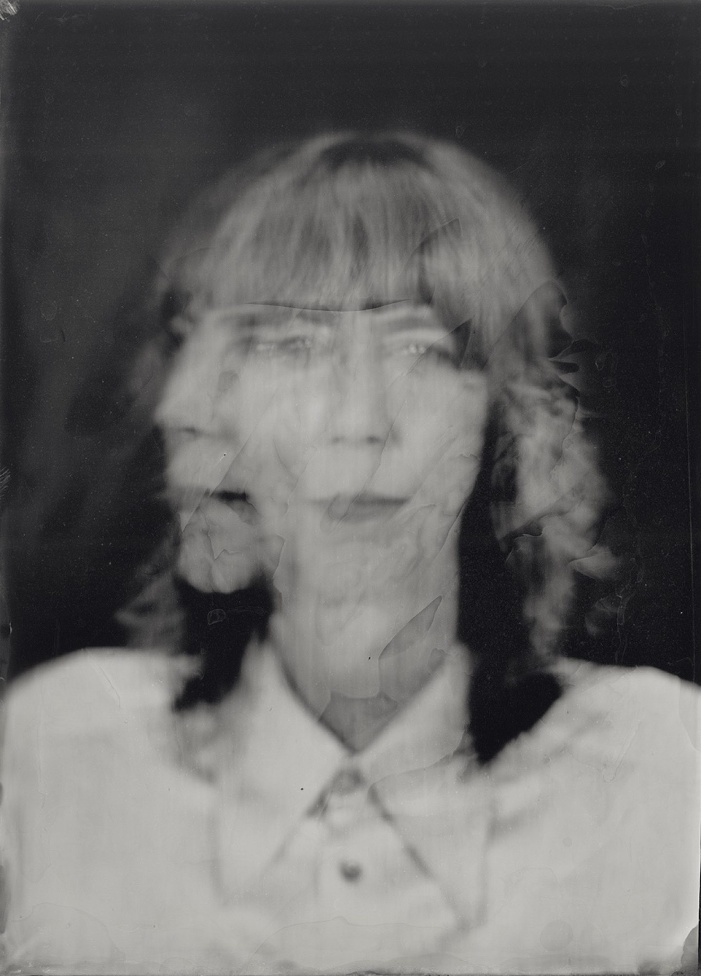 A black-and-white, heavily blurred portrait of a person with braided hair and bangs. The image appears to have multiple overlapping exposures, creating a ghostly, distorted effect where facial features are doubled and smeared. The person seems to be wearing a light-colored shirt with a collar, but details are obscured by the motion blur and damage-like marks across the surface. The overall mood is eerie and abstract, with a vintage, degraded photograph quality.