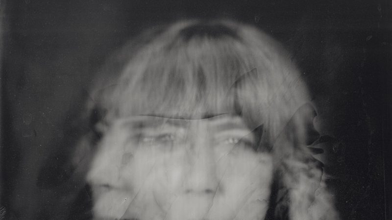 A black-and-white, heavily blurred portrait of a person with braided hair and bangs. The image appears to have multiple overlapping exposures, creating a ghostly, distorted effect where facial features are doubled and smeared. The person seems to be wearing a light-colored shirt with a collar, but details are obscured by the motion blur and damage-like marks across the surface. The overall mood is eerie and abstract, with a vintage, degraded photograph quality.