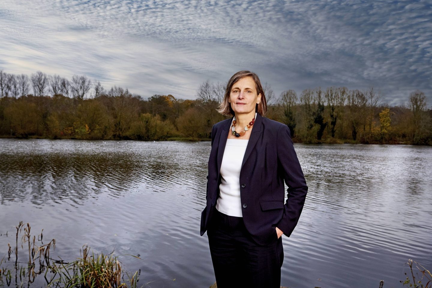 A person in a dark suit stands by a calm lake, with autumn trees and a textured cloudy sky in the background.