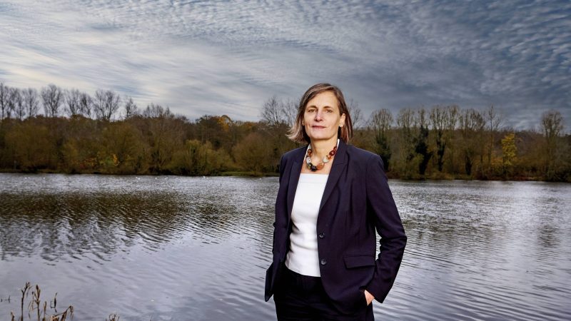A person in a dark suit stands by a calm lake, with autumn trees and a textured cloudy sky in the background.