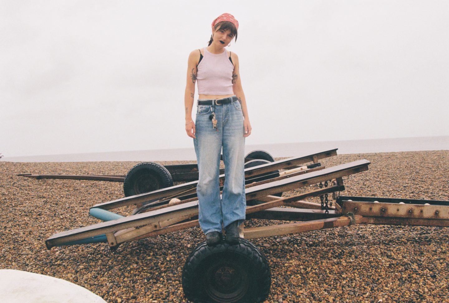 Person standing on a metal trailer frame on a pebble beach, wearing jeans, a pink top, and a bandana.