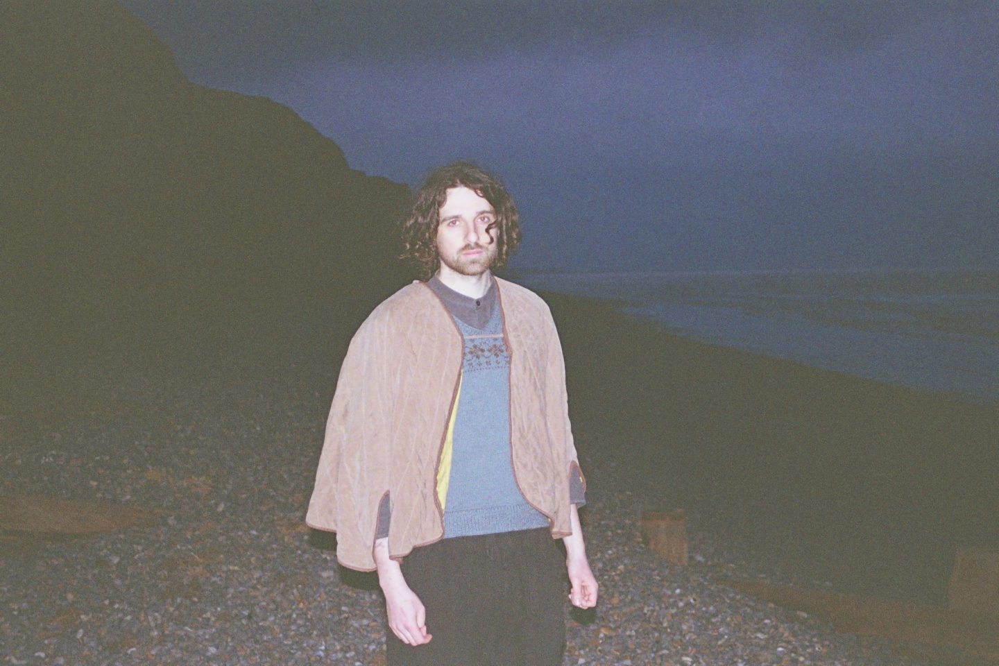 Person standing on a rocky beach at dusk, wearing a sweater and draped shawl, with cliffs on one side and the ocean in the background.