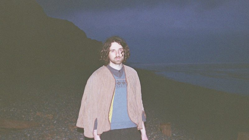 Person standing on a rocky beach at dusk, wearing a sweater and draped shawl, with cliffs on one side and the ocean in the background.
