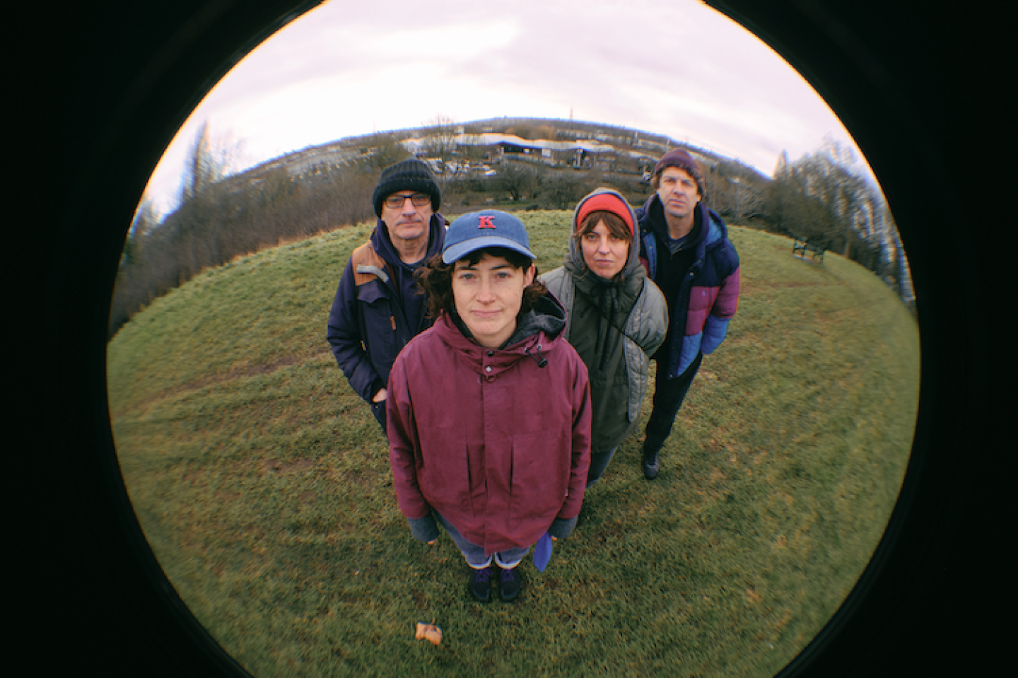 Four people stand close together on a grassy hill, looking up at a fisheye camera, with an overcast sky and distant buildings in the background.