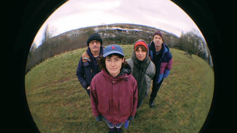 Four people stand close together on a grassy hill, looking up at a fisheye camera, with an overcast sky and distant buildings in the background.