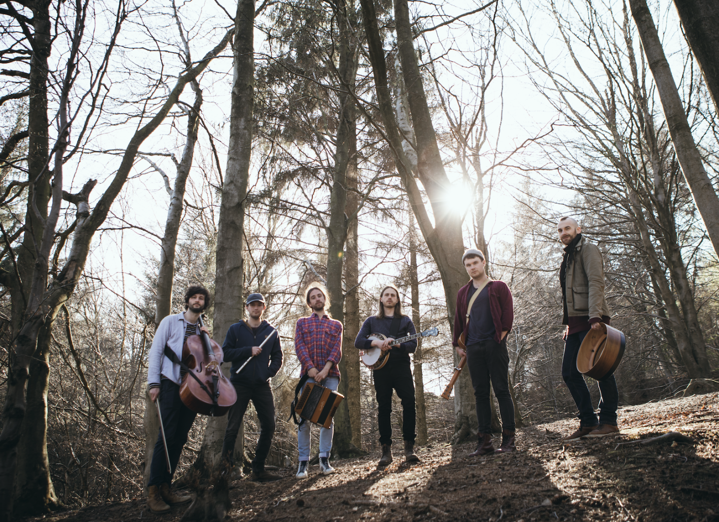 A group of six men stand outdoors in a wooded area, surrounded by tall, leafless trees with sunlight streaming through the branches behind them. They are casually dressed and appear to be a band, each holding or standing near musical instruments. Visible instruments include a cello, a flute, an accordion, a banjo, and a guitar.