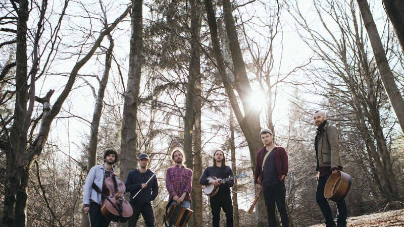 A group of six men stand outdoors in a wooded area, surrounded by tall, leafless trees with sunlight streaming through the branches behind them. They are casually dressed and appear to be a band, each holding or standing near musical instruments. Visible instruments include a cello, a flute, an accordion, a banjo, and a guitar.